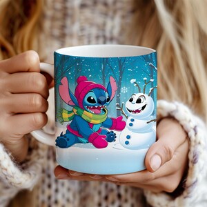 May include: A white ceramic mug featuring Stitch from Lilo & Stitch, wearing a pink hat, scarf, and gloves, and Olaf the snowman. The mug has a winter forest scene background with falling snow. The mug is held by a person.