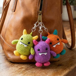 May include: Three colorful crochet monster keychains. The monsters are green, purple, and orange, each with a unique color combination for their horns, feet, and arms. They have white teeth and black eyes. The keychains are attached to a brown leather bag.