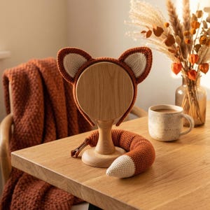 May include: A handcrafted fox ear headband and tail set. The headband features brown and cream-colored crochet fox ears. The matching tail is orange and cream-colored. The set is displayed on a wooden stand on a wooden table.