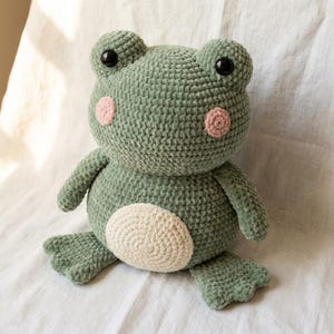 Puede incluir: Un peluche de rana de ganchillo verde hecho a mano con ojos negros y mejillas rosas. La rana tiene una barriga color crema y está sentada sobre una superficie blanca. Este juguete blando es un artículo lindo y tierno.