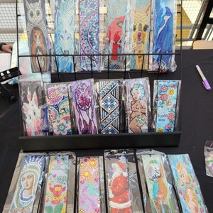 May include: A display of colourful diamond painting bookmarks. The bookmarks feature various designs, including animals, flowers, religious figures, and inspirational quotes.  The bookmarks are displayed on a black wire rack.