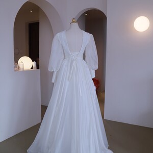 May include: A white wedding dress with a low back and long, sheer sleeves. The dress features a flowing skirt with a subtle floral design near the hem. The back of the dress has a lace-up detail. The dress is displayed on a mannequin.