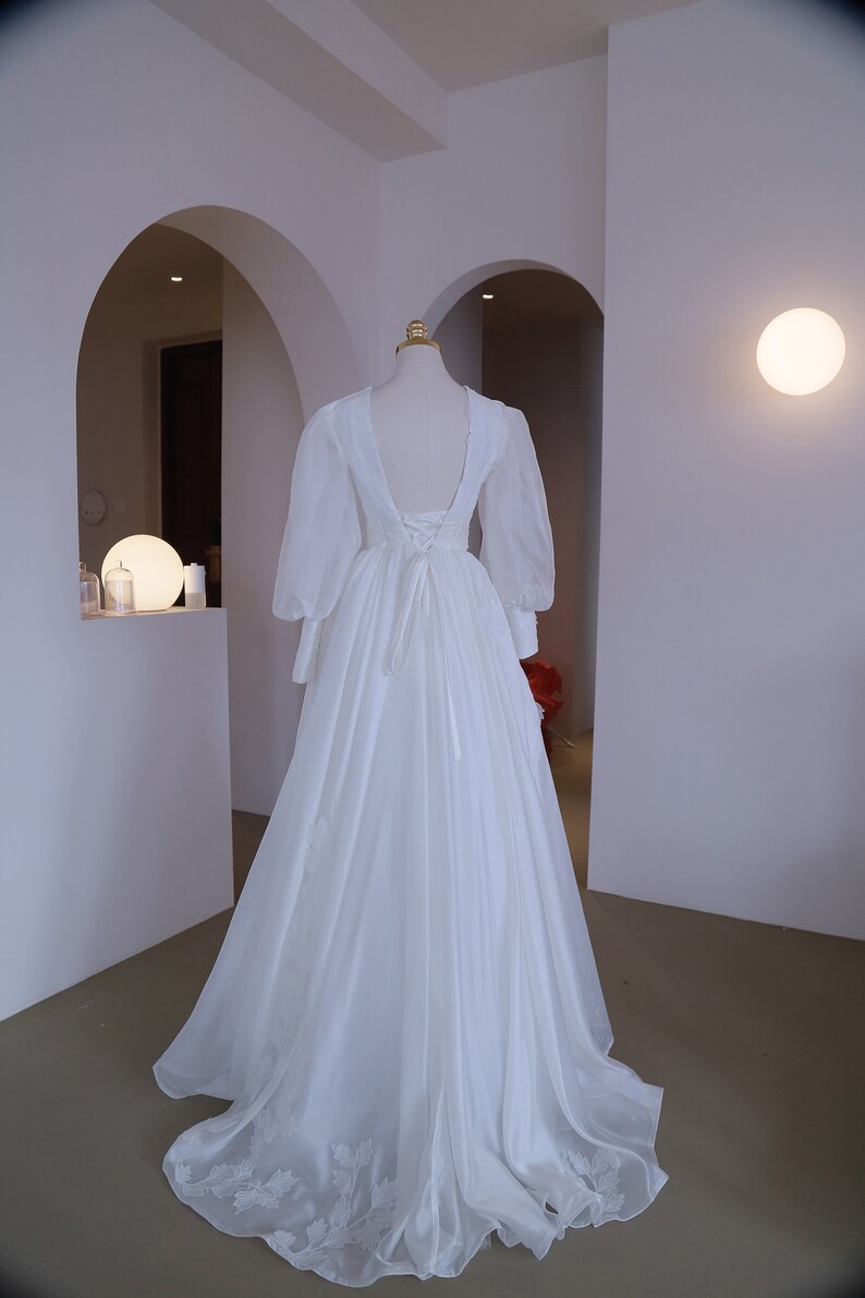 May include: A white wedding dress with a low back and long, sheer sleeves. The dress features a flowing skirt with a subtle floral design near the hem. The back of the dress has a lace-up detail. The dress is displayed on a mannequin.