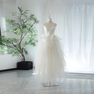 May include: A white strapless wedding dress with a fitted bodice and a flowing tulle skirt. The dress has a large white flower embellishment on the bodice.