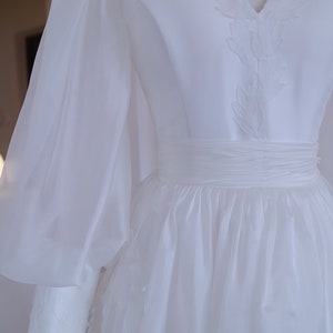 May include: A white wedding dress with a V-neckline and long, sheer sleeves. The bodice features delicate embroidered detailing, and the waist is cinched with a matching sash. The skirt flows gracefully, creating an elegant silhouette.