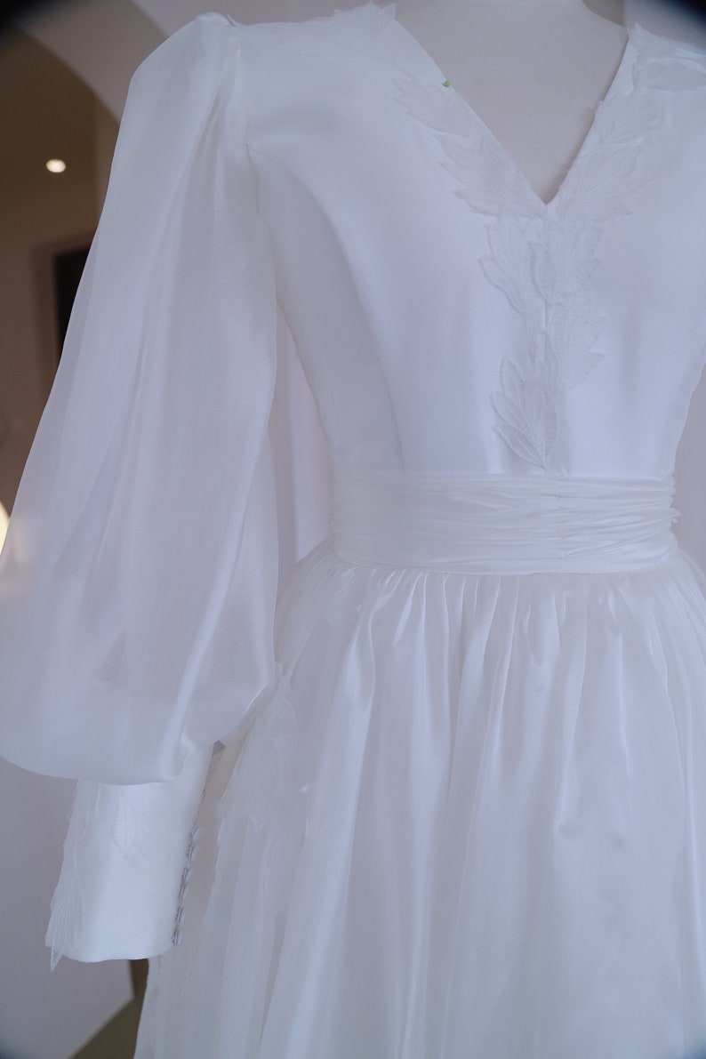 May include: A white wedding dress with a V-neckline and long, sheer sleeves. The bodice features delicate embroidered detailing, and the waist is cinched with a matching sash. The skirt flows gracefully, creating an elegant silhouette.