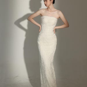 Peut inclure: Une robe ivoire sans bretelles, longue jusqu'au sol, avec des détails de dentelle complexes. La robe a une silhouette ajustée qui s'évase légèrement à l'ourlet. La modèle porte des talons blancs à bout pointu. Le fond est un mur blanc uni.