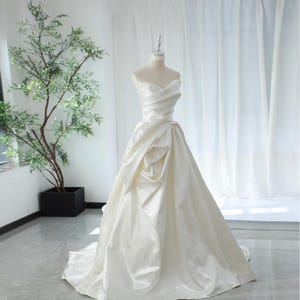 May include: A white strapless satin wedding dress with a fitted bodice and a flowing skirt. The skirt has a gathered design at the waist.