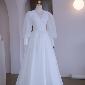 May include: A white wedding dress with a V-neckline and long, sheer, puffy sleeves. The dress features a fitted bodice and a flowing skirt. The dress is displayed on a mannequin in a well-lit room.