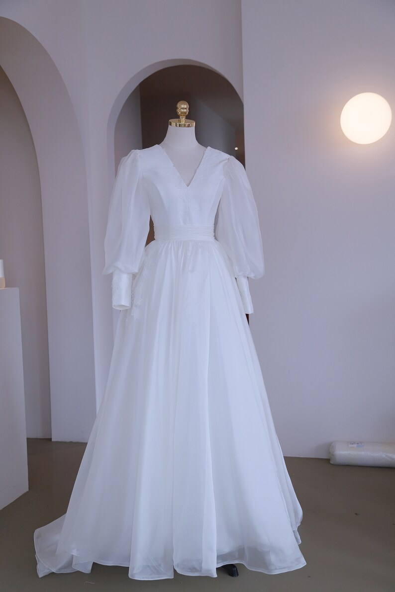 May include: A white wedding dress with a V-neckline and long, sheer, puffy sleeves. The dress features a fitted bodice and a flowing skirt. The dress is displayed on a mannequin in a well-lit room.
