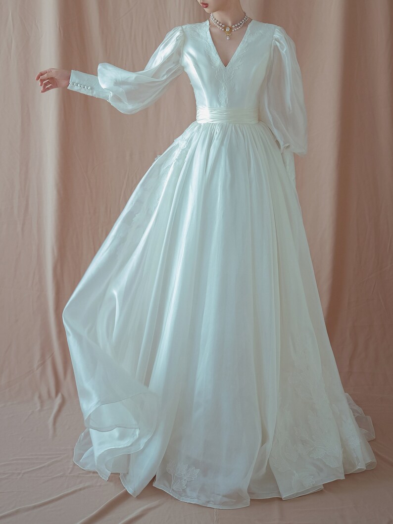 May include: A white wedding dress with a V-neckline and long, sheer sleeves. The dress features a fitted bodice, a defined waist, and a flowing skirt. Delicate floral embroidery adorns the bodice and skirt. The dress is made of a lightweight, flowing fabric.
