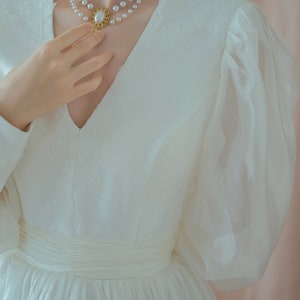 May include: A white dress with a V-neckline and long, sheer sleeves. The dress features a cinched waist and a layered skirt. A double-strand pearl necklace with a gold pendant is worn around the neck. The dress has floral embroidery.
