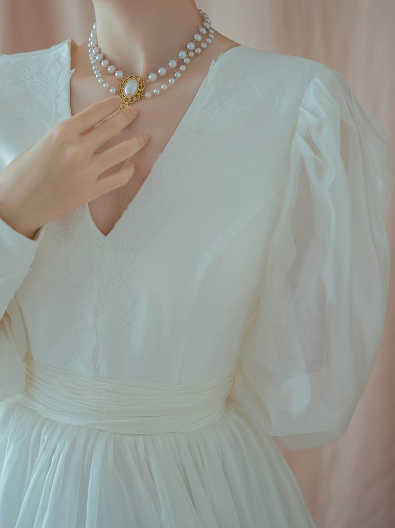 May include: A white dress with a V-neckline and long, sheer sleeves. The dress features a cinched waist and a layered skirt. A double-strand pearl necklace with a gold pendant is worn around the neck. The dress has floral embroidery.
