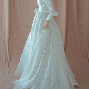 May include: A white wedding dress with a V-neckline and long, puffy sleeves. The dress features a fitted bodice and a flowing skirt with floral embroidery near the hem. The fabric appears to be a lightweight, possibly silk or chiffon.