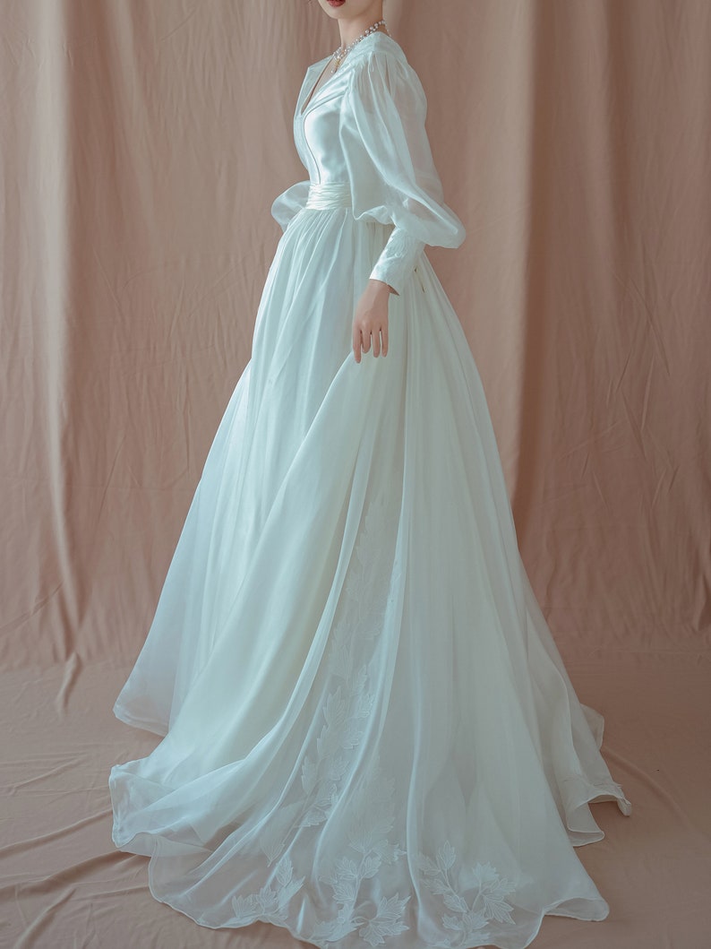 May include: A white wedding dress with a V-neckline and long, puffy sleeves. The dress features a fitted bodice and a flowing skirt with floral embroidery near the hem. The fabric appears to be a lightweight, possibly silk or chiffon.