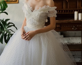 Pode incluir: Um vestido de noiva branco de ombros descobertos com um corpete de renda e uma saia de tule com bolinhas brancas. O vestido é usado por uma modelo em pé em frente a um armário de madeira.