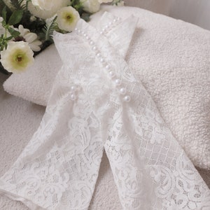 Puede incluir: Elegantes guantes de encaje blanco con detalles de perlas. Los guantes están adornados con delicados diseños florales y una fila de perlas en la parte superior. Un ramo de flores blancas descansa en el fondo, creando una estética suave y romántica.