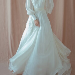 May include: A white wedding dress with a V-neckline and long, puffy sleeves. The dress features delicate embroidery on the bodice and a flowing skirt. The fabric appears to be a lightweight material, creating a soft, ethereal look. The dress is floor-length.