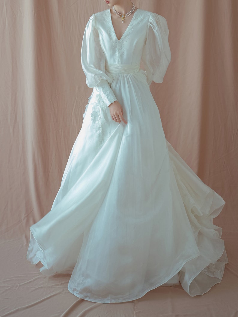 May include: A white wedding dress with a V-neckline and long, puffy sleeves. The dress features delicate embroidery on the bodice and a flowing skirt. The fabric appears to be a lightweight material, creating a soft, ethereal look. The dress is floor-length.