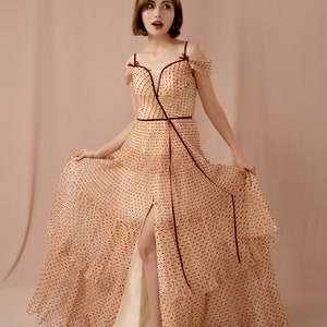 May include: A woman wearing a beige dress with red polka dots and a burgundy ribbon. The dress has a V-neckline, off-the-shoulder straps, and a thigh-high slit. The dress is made of a sheer fabric and has a ruffled hem.