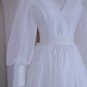 May include: A white wedding dress with a V-neckline and long, sheer sleeves. The bodice features delicate embroidered details, and the waist is cinched with a matching sash. The skirt flows gracefully, creating an elegant silhouette.