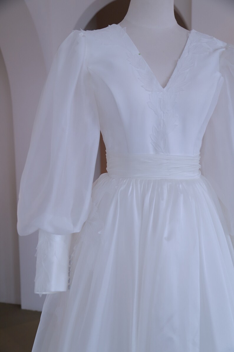 May include: A white wedding dress with a V-neckline and long, sheer sleeves. The bodice features delicate embroidered details, and the waist is cinched with a matching sash. The skirt flows gracefully, creating an elegant silhouette.