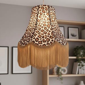 Può includere: Un paralume con motivo leopardato marrone e beige. Il paralume ha un bordo smerlato con lunghe frange beige. È appeso a un cavo bianco.