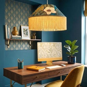 Può includere: Un ufficio domestico con una scrivania in legno, un computer e una sedia gialla. Un paralume a motivi floreali con frange è appeso sopra. La stanza ha pareti blu e carta da parati a motivi.