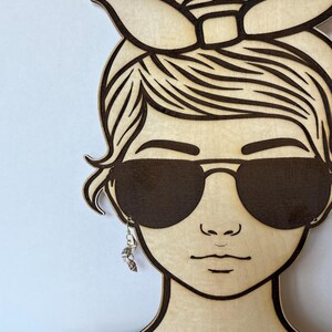 May include: Wooden earring display in the shape of a woman's face wearing sunglasses and a headscarf. The display holds a pair of dangling earrings with a seashell design. The wood has a light, natural finish with dark brown etched details.