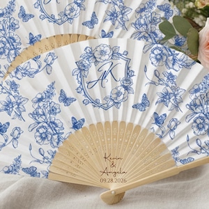 以下が含まれることがあります： 白い背景に青い花と蝶の模様が描かれた2つの折りたたみ式扇子。各扇子の中央にはAKのイニシャルが入った紋章があります。手前の扇子の木製の骨組みには、Kevin & Angelaという文字と2026年9月28日という日付が刻印されています。