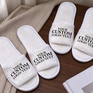 May include: Two pairs of white plush open-toe slippers featuring a soft, fuzzy texture. Each slipper is printed with the black text 'YOUR CUSTOM LOGO/TEXT' on the top strap. The slippers have a simple slip-on design with a white sole and lining.