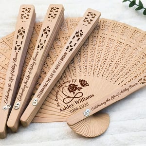 May include: A set of wooden hand fans with a laser-cut design. The fans are personalized with the text "Celebrating the Life of Ashley Brown" and "In Loving Memory of Ashley Williams 1984-2025".