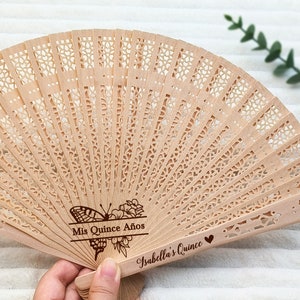 Puede incluir: Un abanico de mano de madera con un dise&ntilde;o delicado e intrincado. El abanico est&aacute; decorado con un motivo de mariposa y flores y el texto "Mis Quince A&ntilde;os Isabella's Quince".