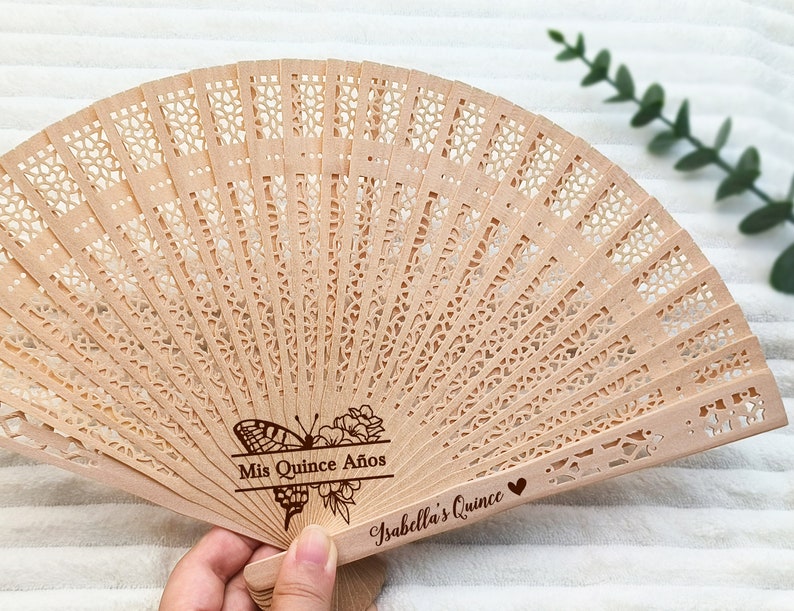 Puede incluir: Un abanico de mano de madera con un dise&ntilde;o delicado e intrincado. El abanico est&aacute; decorado con un motivo de mariposa y flores y el texto "Mis Quince A&ntilde;os Isabella's Quince".