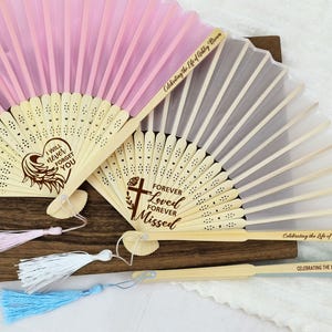 May include: Two wooden folding fans with pink and white silk fabric. The fans are decorated with laser-engraved messages, "I will never forget you" and "Forever Loved Forever Missed". The fans have tassels in pink, white, and blue.