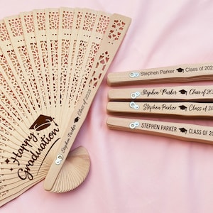 May include: Wooden graduation fans with intricate cut-out designs. One fan is open, displaying "Happy Graduation" and a graduation cap graphic. The handles of the fans are engraved with "Stephen Parker Class of 2025".