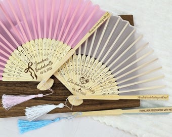 May include: Pink and white folding fans with wooden handles and decorative tassels. The fans are personalized with text that reads "Sandra's Bachelorette" and "Thanks for celebrating with me!" on the handles. The fans are on a wooden surface.