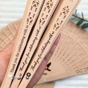 May include: Three wooden hand fans with intricate cutouts and the text "Thank you for celebrating with us" engraved on each. The fans are closed and held in a hand.