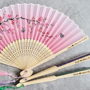 Può includere: Tre ventagli rosa e bianchi con un motivo a fiori di ciliegio e un manico in legno. I ventagli sono personalizzati con i nomi "Sarah Adam" e la data "06.18.2025".