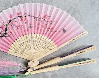 May include: Three pink and white hand fans with a cherry blossom design and a wooden handle. The fans have personalized text with the names "Sarah Adam" and the date "06.18.2025".