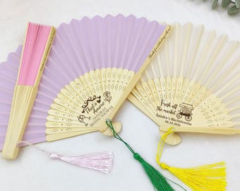 May include: A collection of decorative folding fans in various colors. One fan is pink, another is lavender, and a third is off-white. Each fan has a wooden handle and is adorned with tassels. Text on the fans includes phrases like "That's Amore" and "Fresh off the market."