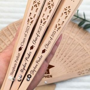 May include: Three wooden fans with intricate cut-out designs are held in a hand. Each fan is engraved with "Stephen Parker" and "Class of 2025" along with a graduation cap icon. A fourth fan is partially visible in the background.