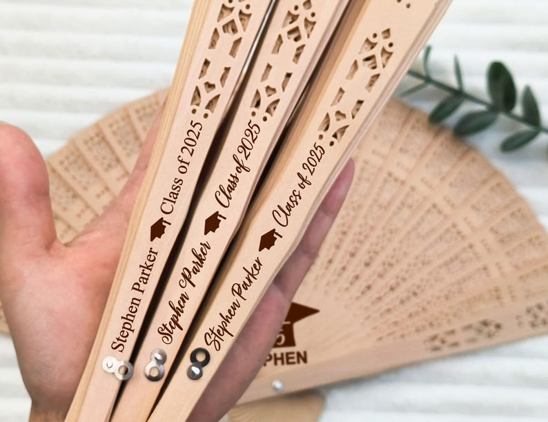 May include: Three wooden fans with intricate cut-out designs are held in a hand. Each fan is engraved with "Stephen Parker" and "Class of 2025" along with a graduation cap icon. A fourth fan is partially visible in the background.