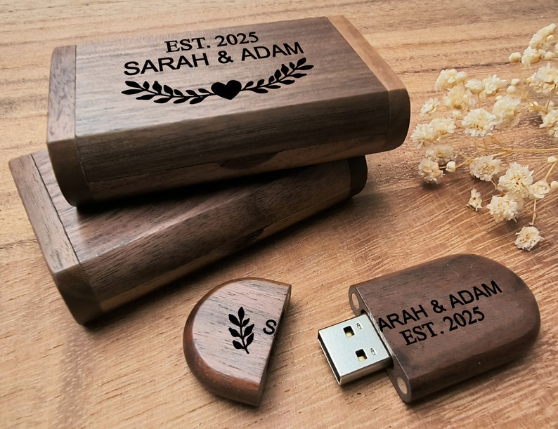 Op de afbeelding: Een houten USB-stick met een gepersonaliseerde inscriptie: "SARAH & ADAM EST. 2025". De stick heeft een rechthoekige vorm met afgeronde hoeken en een donkerbruine afwerking. De inscriptie is gegraveerd in wit. De stick wordt geleverd met een houten doos met een vergelijkbare inscriptie en ontwerp.
