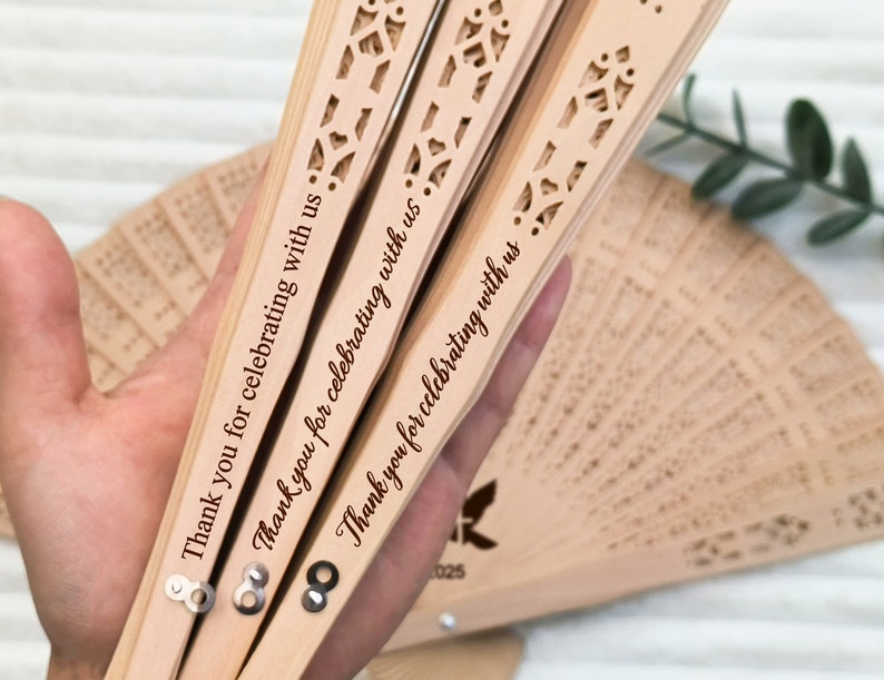 May include: Three wooden hand fans with intricate cutouts and the text "Thank you for celebrating with us" engraved on each. The fans are closed and held in a hand.