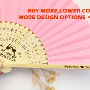 May include: Pink paper folding fan with a wooden handle. The fan has a brown design with the text "2025 GRADUATE" and two wine glasses clinking. The handle has the text "Stephen Parker Class of 2025".