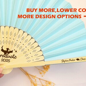 May include: A light blue hand fan with a wooden handle and a graduation design. The fan has a white and gold pattern and the text "Graduate 2025" is printed on the handle. The text "Stephen Parker Class of 2025" is printed on the back of the handle.