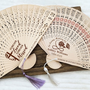 May include: Two wooden hand fans with intricate cutouts. One fan has a floral design and the text "In Loving Memory of Ashley Williams 1984-2025 Celebrating the Life of Ashley Brown". The other fan has a star and moon design and the text "Forever Loved Forever Missed Celebrating the Life of Ashley Brown".