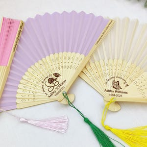 以下が含まれることがあります： 木製の枠と布製の羽根を持つ手持ち扇子が3つあります。扇子はレーザー彫刻された文字とデザインで飾られています。1つの扇子はピンク、1つはラベンダー、もう1つは白です。扇子に書かれた文字は、「Celebrating the life of Ashley」、「In loving memory of Ashley Williams 1984-2025」、「In loving memory of Ashley Williams 1984-2025」です。