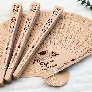 May include: Several wooden fans with intricate cut-out designs are displayed. The fans are engraved with "Stephen Parker Class of 2025" and a graduation cap. The fans are light brown in color and appear to be made of wood.